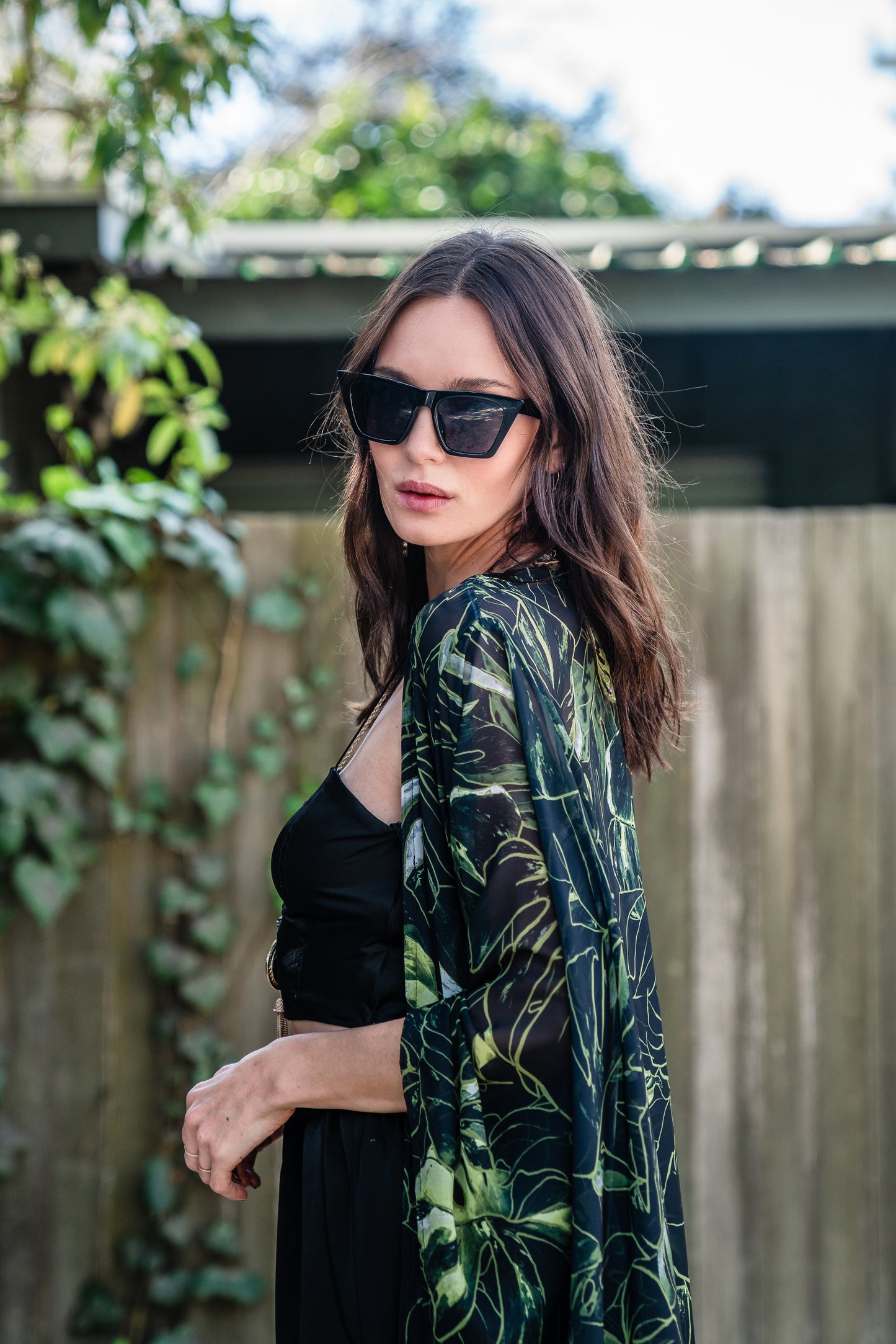 Model wears black chiffon Kimono with varying green palm leaf print. The kimono has classic wide kimono sleeves and lands at midi length. Kimono has a matching tie belt.