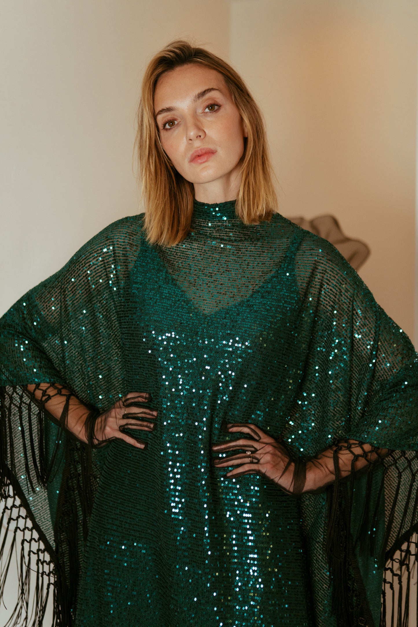 This caftan is a dazzling, full-length garment featuring a shimmering jade green sequin overlay on a mesh fabric. The sequins catch the light, creating a sparkling effect throughout. It has a mock neck collar with a straight, loose fit that drapes elegantly over the body. The edges of the caftan, including the hem and the sides, are embellished with long, silk black fringe that add movement and a playful flair.