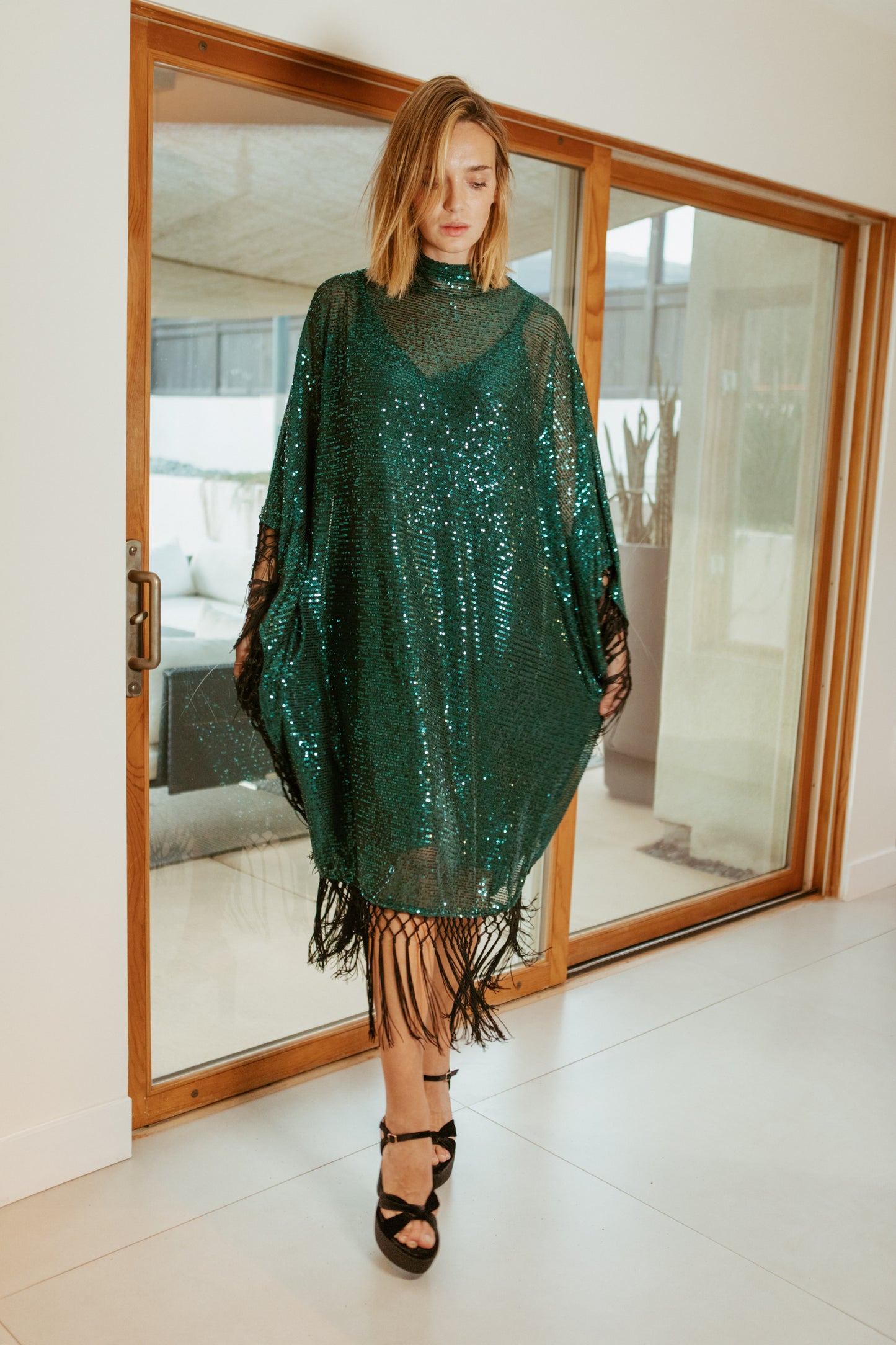 This caftan is a dazzling, full-length garment featuring a shimmering jade green sequin overlay on a mesh fabric. The sequins catch the light, creating a sparkling effect throughout. It has a mock neck collar with a straight, loose fit that drapes elegantly over the body. The edges of the caftan, including the hem and the sides, are embellished with long, silk black fringe that add movement and a playful flair.