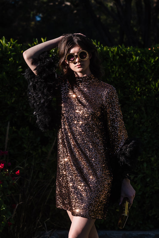 This shimmering copper flash mod mini dress has it all - glittering sequins, delicate mock neck, and long bell sleeves cuffed with black feathers. Retro 60s glam