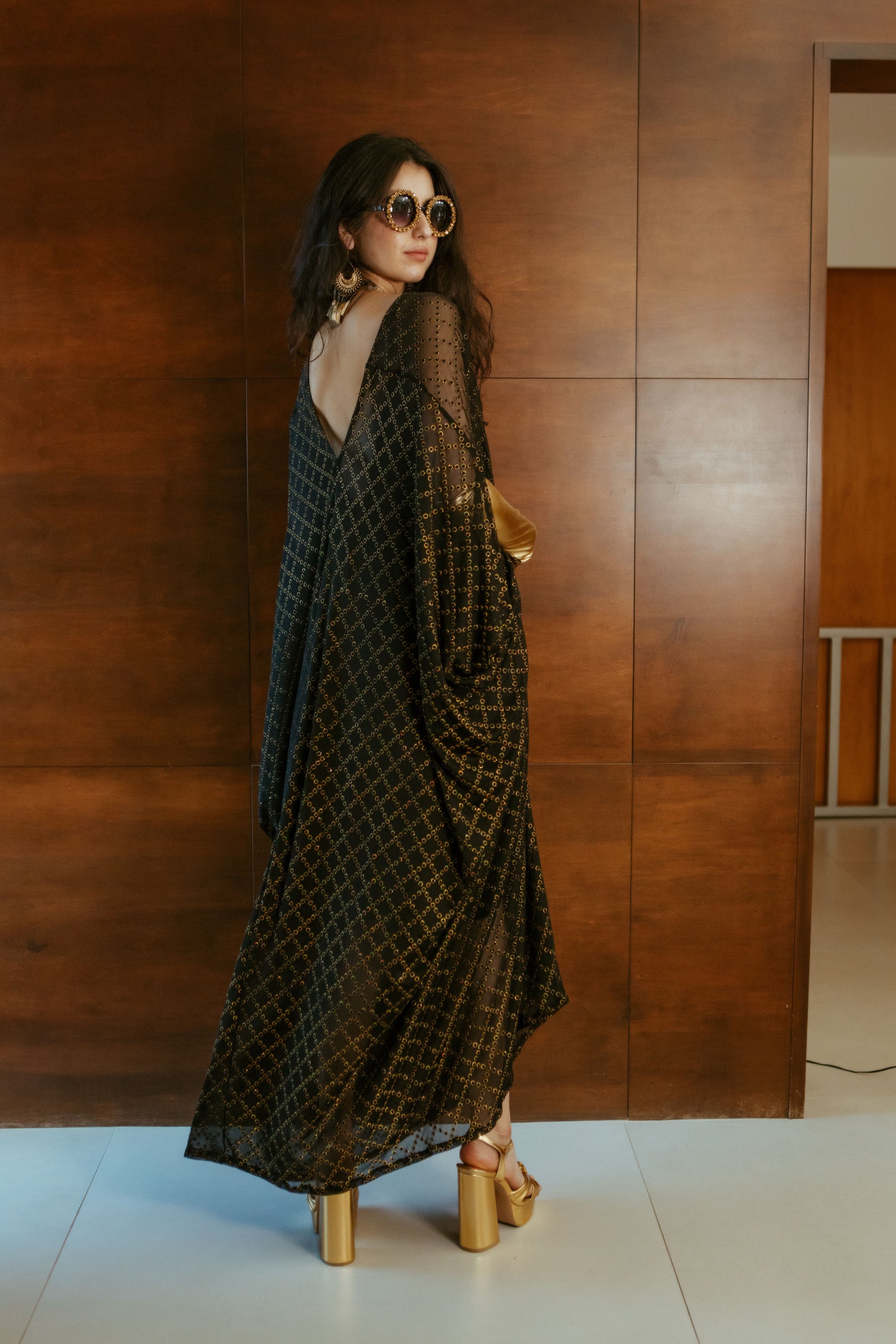 Black, sheer mesh caftan dress embellished with shimmering metallic gold circles in a geometric pattern that creates a sort of chain-link effect. Featuring a deep v-neckline, short batwing sleeves, and an ankle-length hem. This caftan is a voluminous garment that gives a flowy silhouette that drapes beautifully on all shapes.