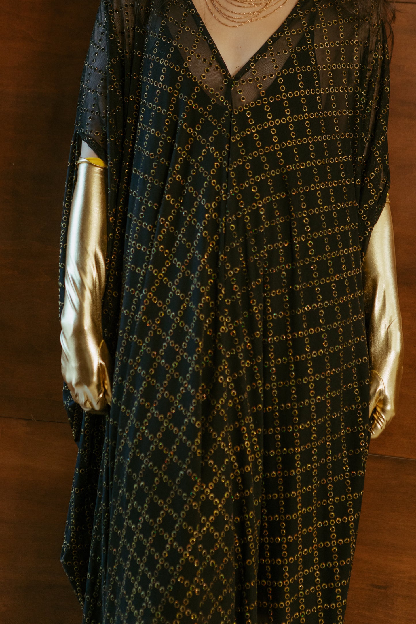 Black, sheer mesh caftan dress embellished with shimmering metallic gold circles in a geometric pattern that creates a sort of chain-link effect. Featuring a deep v-neckline, short batwing sleeves, and an ankle-length hem. This caftan is a voluminous garment that gives a flowy silhouette that drapes beautifully on all shapes.