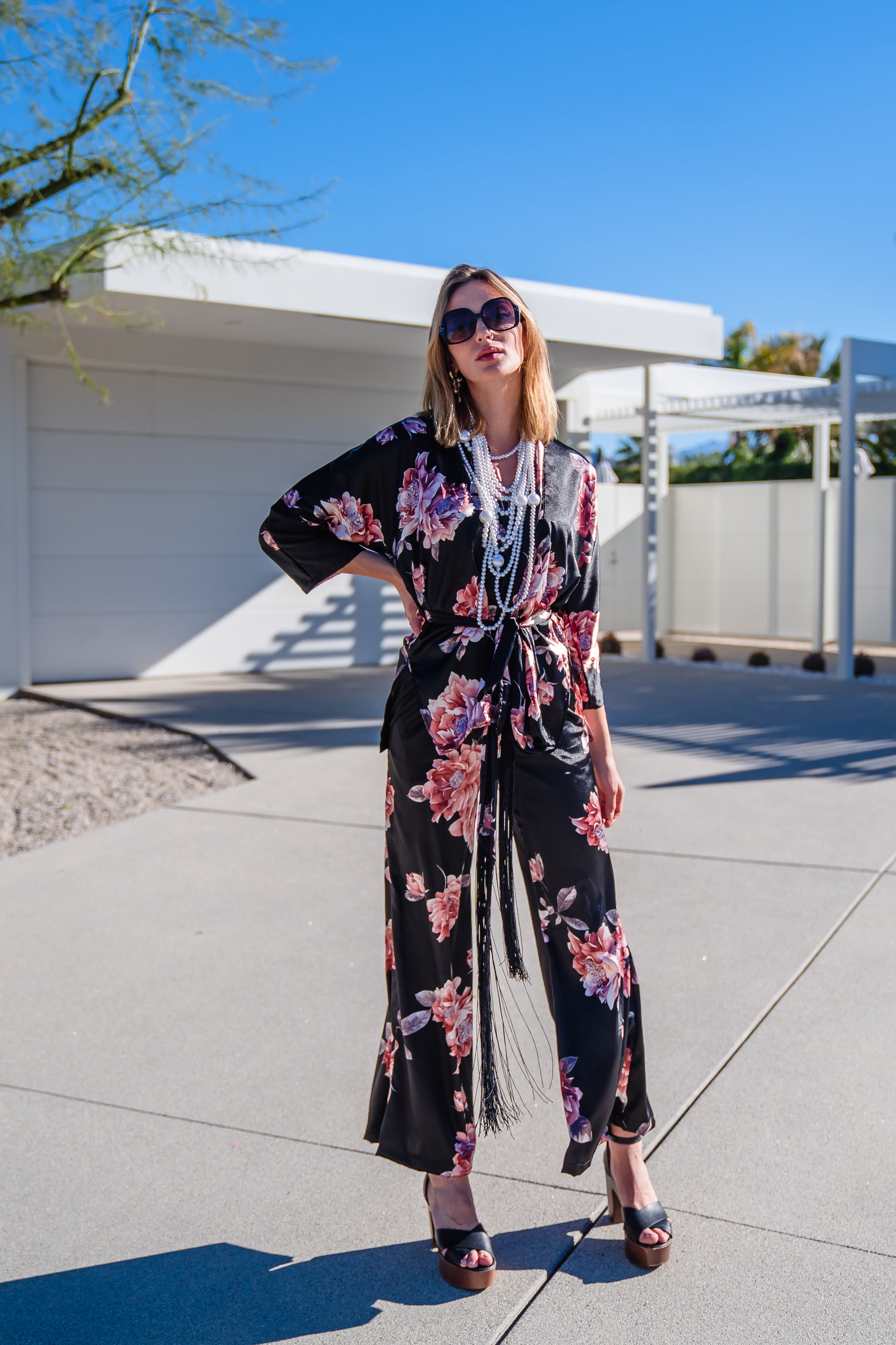 Loungewear set in dark floral pattern. A black background with light pink flowers, this set comes with a wrap dolman jacket.  Sleeves have a forearm hem, and it can be worn as an open jacket or wrap with matching tie belt. The belt features fringe tassels. The matching pants have pockets and an ankle length hem. 