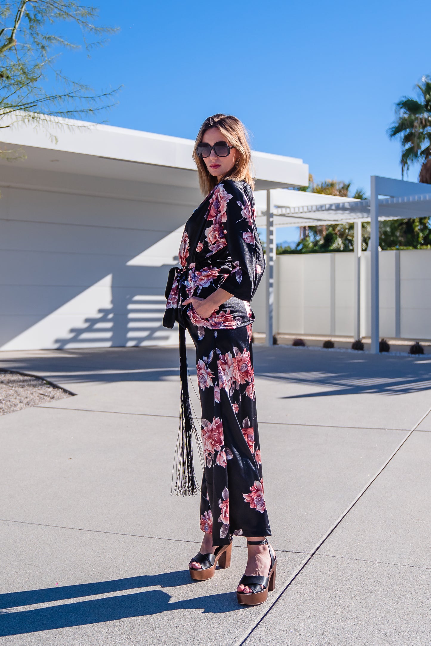 Loungewear set in dark floral pattern. A black background with light pink flowers, this set comes with a wrap dolman jacket.  Sleeves have a forearm hem, and it can be worn as an open jacket or wrap with matching tie belt. The belt features fringe tassels. The matching pants have pockets and an ankle length hem. 