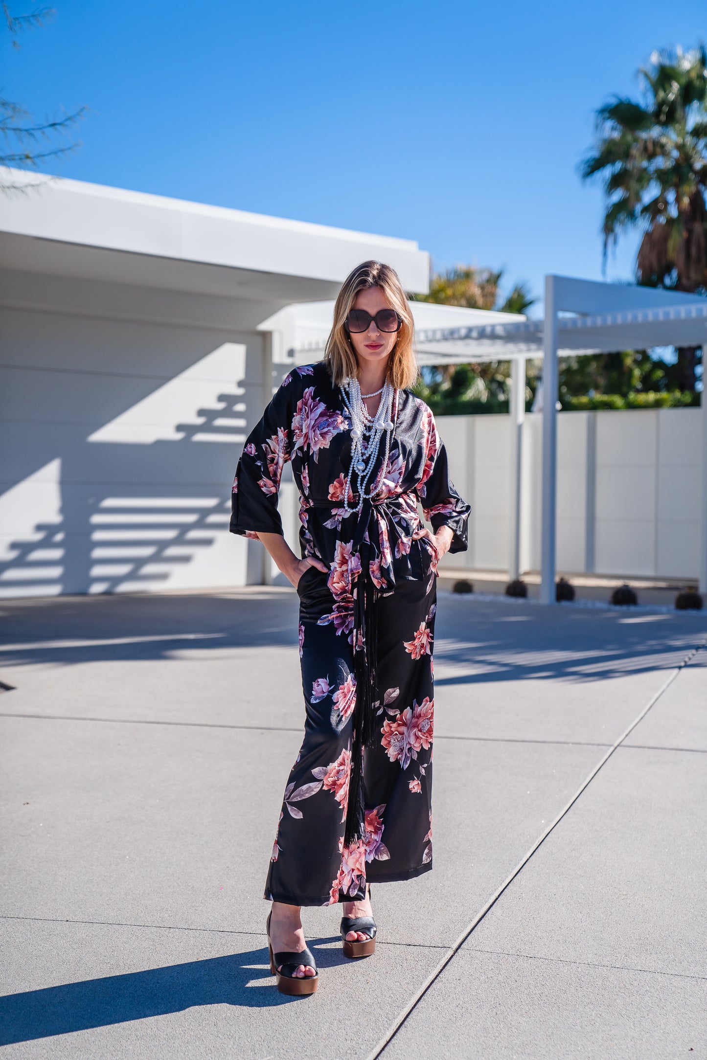 Loungewear set in dark floral pattern. A black background with light pink flowers, this set comes with a wrap dolman jacket.  Sleeves have a forearm hem, and it can be worn as an open jacket or wrap with matching tie belt. The belt features fringe tassels. The matching pants have pockets and an ankle length hem. 