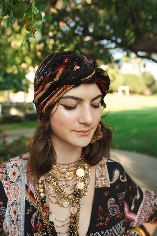 Black mesh fashion turban embellished with an abstract pattern of black, red, and gold sequins. Wears similar to a beanie, with ruching of the fabric that meets front and center at an elegant knot. Vintage-inspired retro 1920s mixed with a modern fabric.