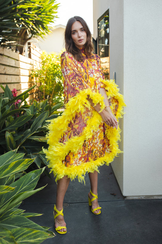 The Intra Dimensiona Mockneck Feather Caftan with slip. A vibrant, fiery abstract print featuring reds and oranges and yellows printed on a semi-sheer, super soft mesh with canary yellow feather border along sleeves and hem. A bohemian dress inspired by Old Hollywood and Palm Springs.