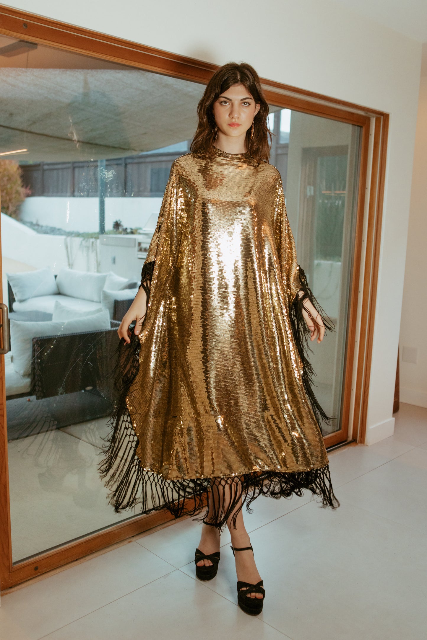 This caftan is a dazzling, full-length garment featuring a shimmering gold sequin overlay on a mesh fabric. The sequins catch the light, creating a sparkling effect throughout. It has a mock neck collar with a straight, loose fit that drapes elegantly over the body. The edges of the caftan, including the hem and the sides, are embellished with long, silk black fringe that add movement and a playful flair to the garment.