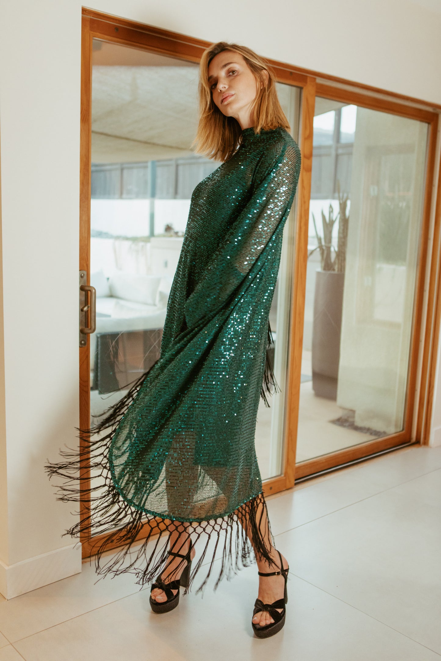 This caftan is a dazzling, full-length garment featuring a shimmering jade green sequin overlay on a mesh fabric. The sequins catch the light, creating a sparkling effect throughout. It has a mock neck collar with a straight, loose fit that drapes elegantly over the body. The edges of the caftan, including the hem and the sides, are embellished with long, silk black fringe that add movement and a playful flair.