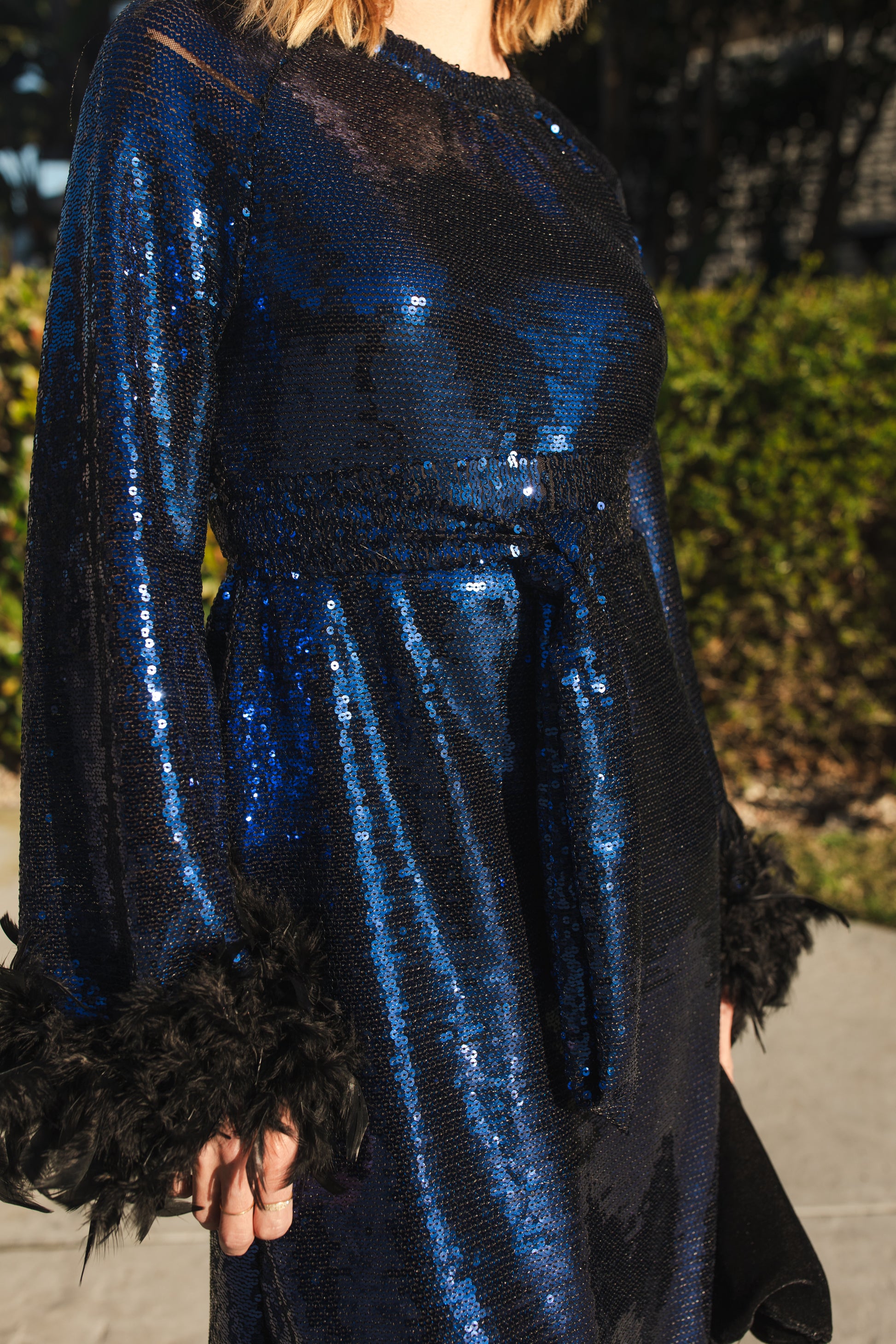 Model wears a knee length dress covered in midnight blue sequins, the dress has a crew neck and full long sleeves with a feather trim.