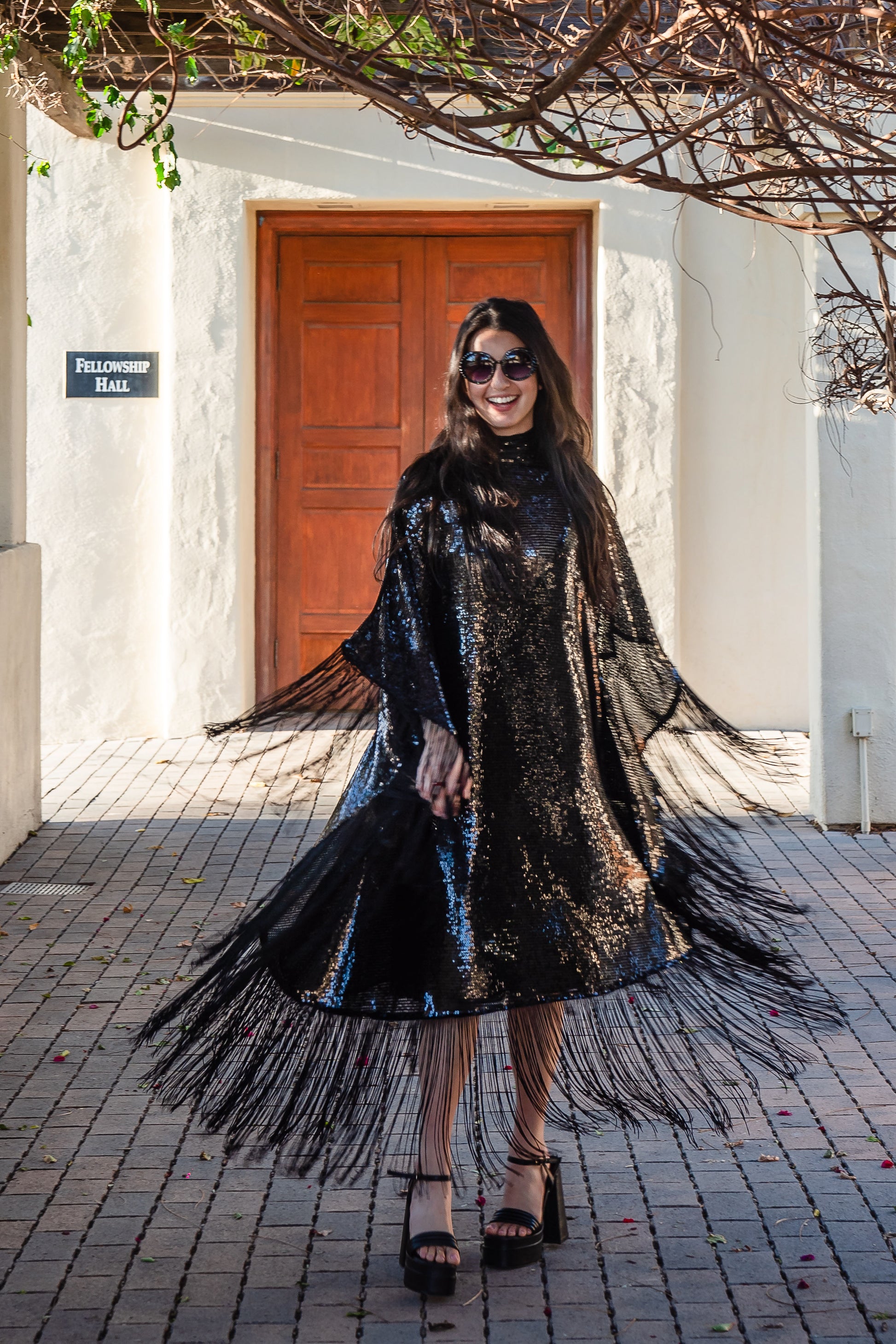 This caftan is a dazzling, full-length garment featuring a shimmering inky black sequin overlay on a mesh fabric. The sequins catch the light, creating a sparkling effect throughout. It has a mock neck collar with a straight, loose fit that drapes elegantly over the body. The edges of the caftan, including the hem and the sides, are embellished with long, black silk fringe that add movement and a playful flair to an elegant and timeless design.