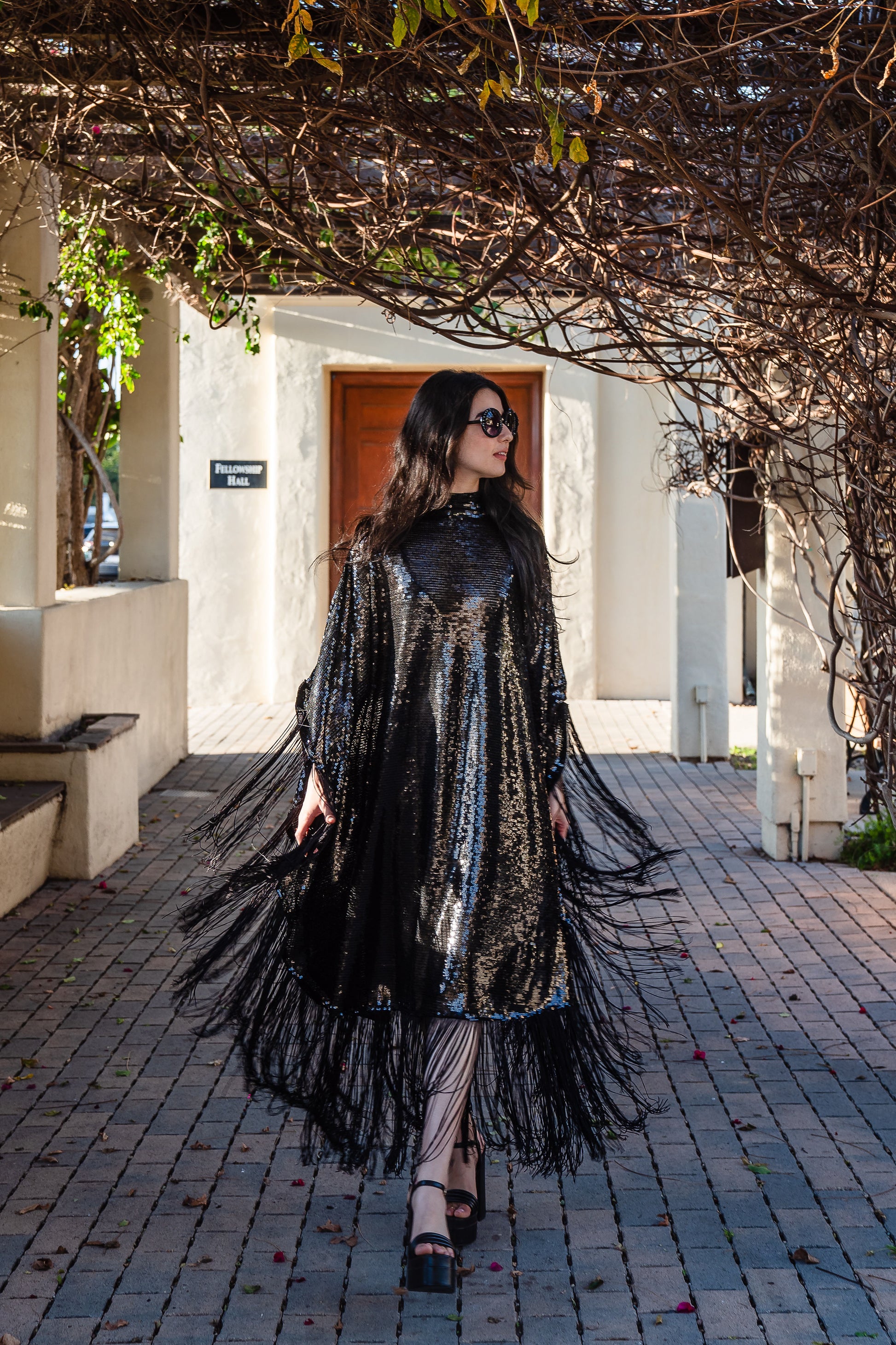 This caftan is a dazzling, full-length garment featuring a shimmering inky black sequin overlay on a mesh fabric. The sequins catch the light, creating a sparkling effect throughout. It has a mock neck collar with a straight, loose fit that drapes elegantly over the body. The edges of the caftan, including the hem and the sides, are embellished with long, black silk fringe that add movement and a playful flair to an elegant and timeless design.
