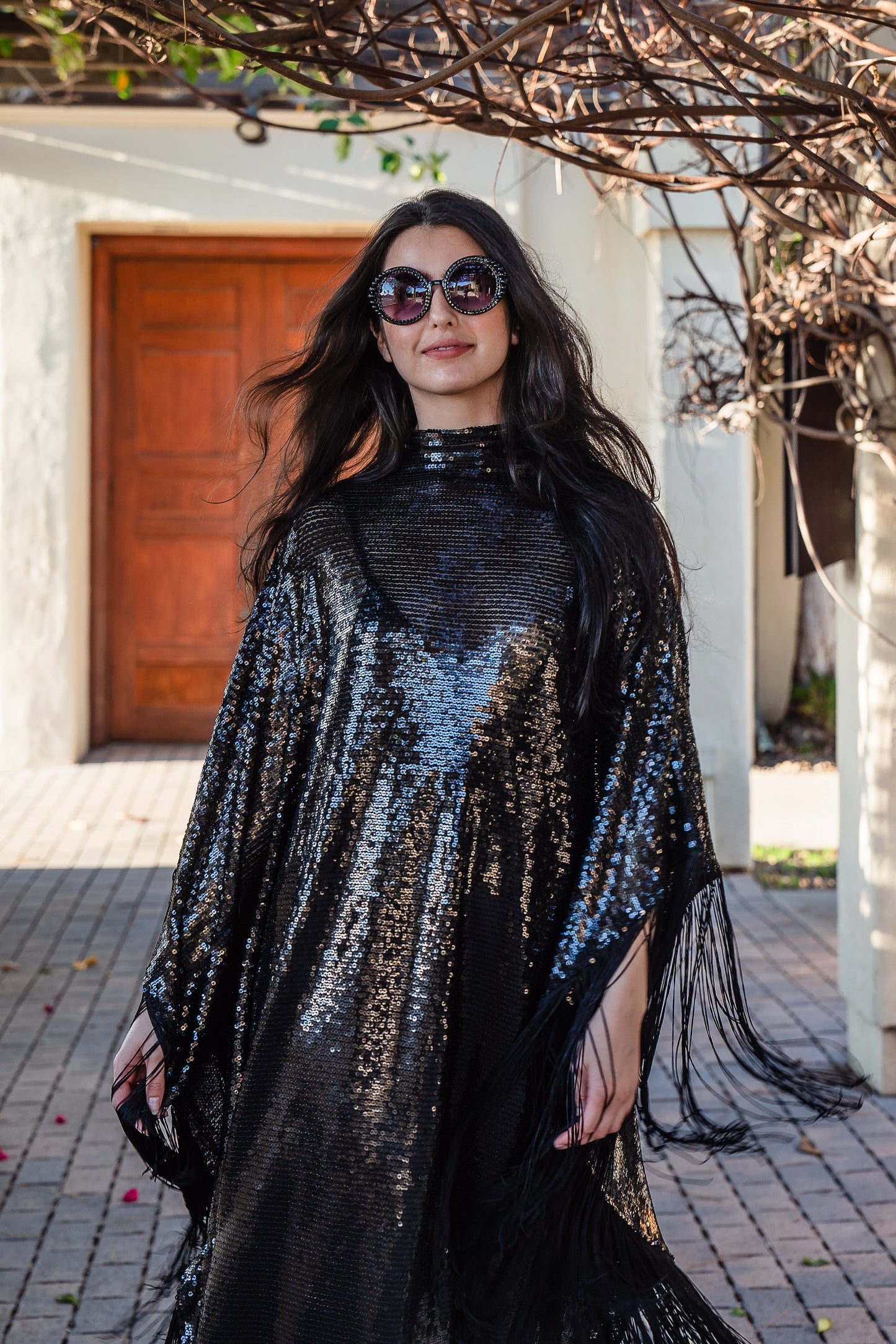 This caftan is a dazzling, full-length garment featuring a shimmering inky black sequin overlay on a mesh fabric. The sequins catch the light, creating a sparkling effect throughout. It has a mock neck collar with a straight, loose fit that drapes elegantly over the body. The edges of the caftan, including the hem and the sides, are embellished with long, black silk fringe that add movement and a playful flair to an elegant and timeless design.