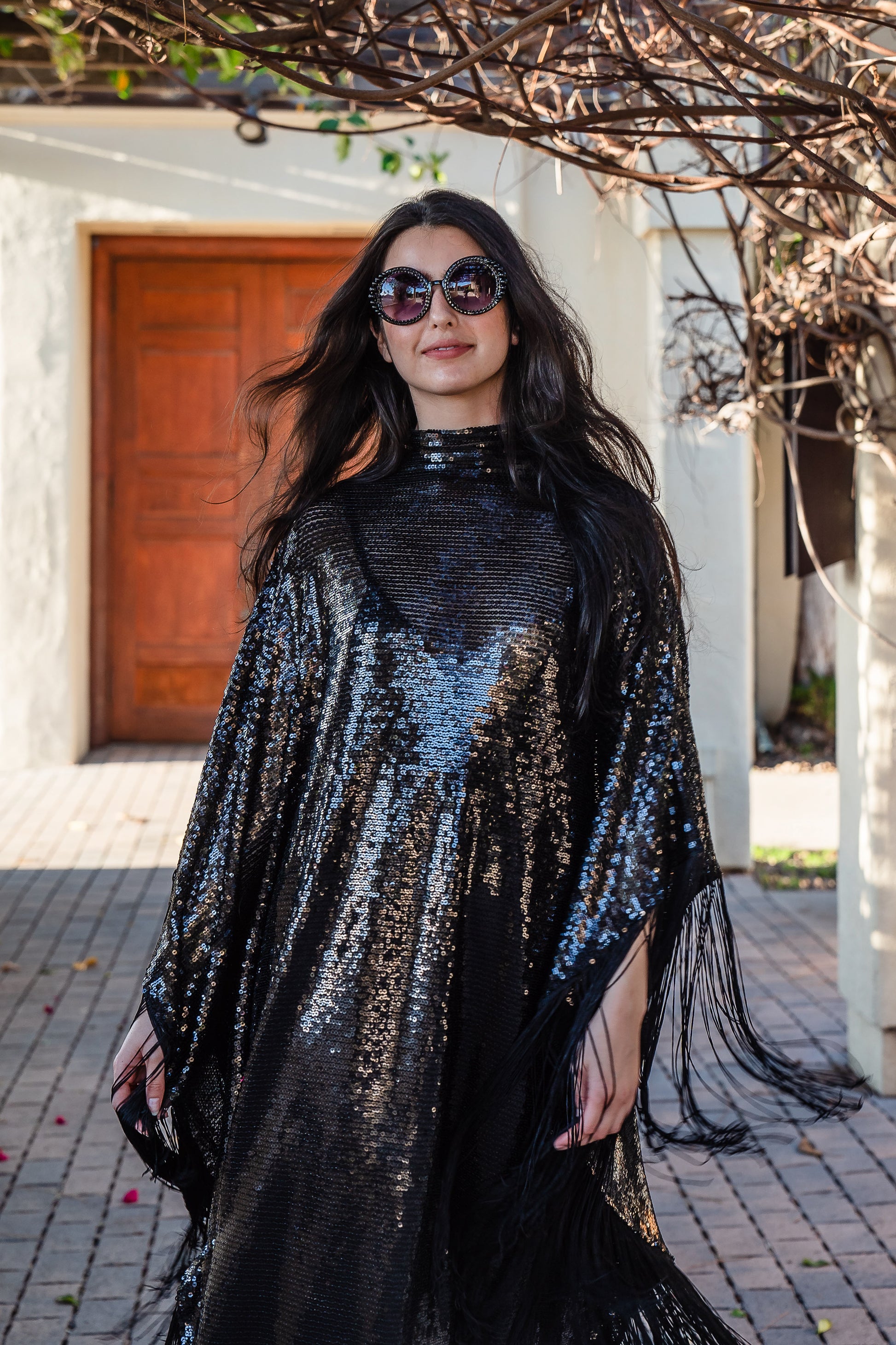 This caftan is a dazzling, full-length garment featuring a shimmering inky black sequin overlay on a mesh fabric. The sequins catch the light, creating a sparkling effect throughout. It has a mock neck collar with a straight, loose fit that drapes elegantly over the body. The edges of the caftan, including the hem and the sides, are embellished with long, black silk fringe that add movement and a playful flair to an elegant and timeless design.