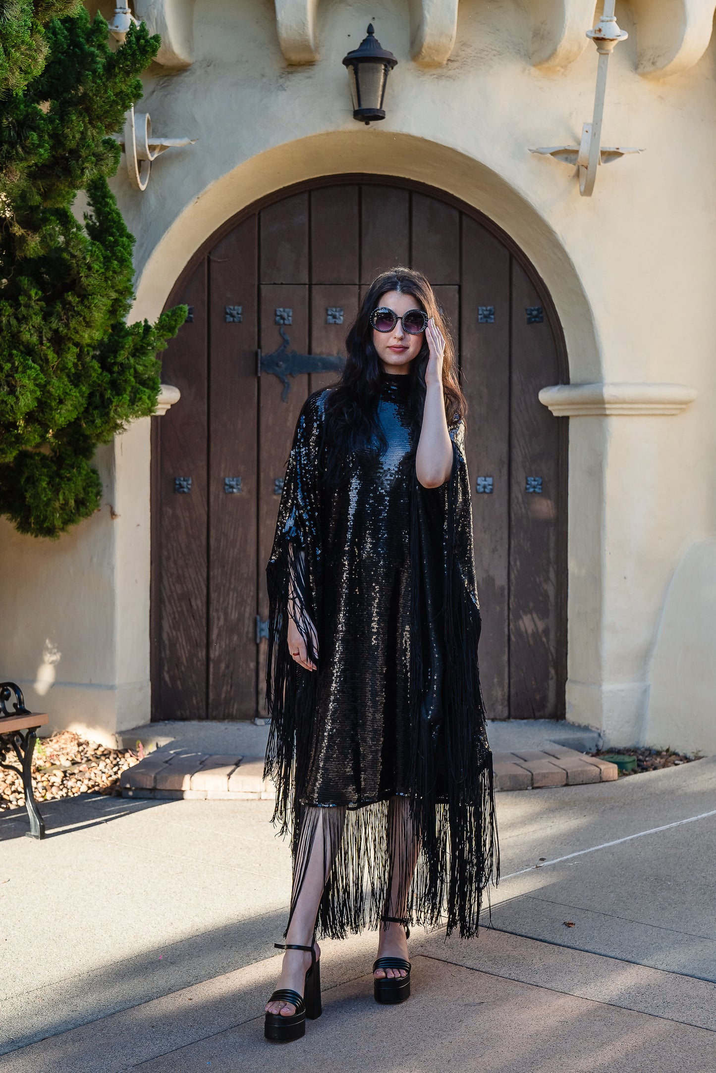 This caftan is a dazzling, full-length garment featuring a shimmering inky black sequin overlay on a mesh fabric. The sequins catch the light, creating a sparkling effect throughout. It has a mock neck collar with a straight, loose fit that drapes elegantly over the body. The edges of the caftan, including the hem and the sides, are embellished with long, black silk fringe that add movement and a playful flair to an elegant and timeless design.