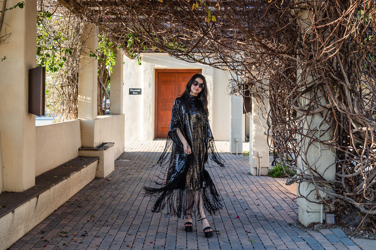 This caftan is a dazzling, full-length garment featuring a shimmering inky black sequin overlay on a mesh fabric. The sequins catch the light, creating a sparkling effect throughout. It has a mock neck collar with a straight, loose fit that drapes elegantly over the body. The edges of the caftan, including the hem and the sides, are embellished with long, black silk fringe that add movement and a playful flair to an elegant and timeless design.