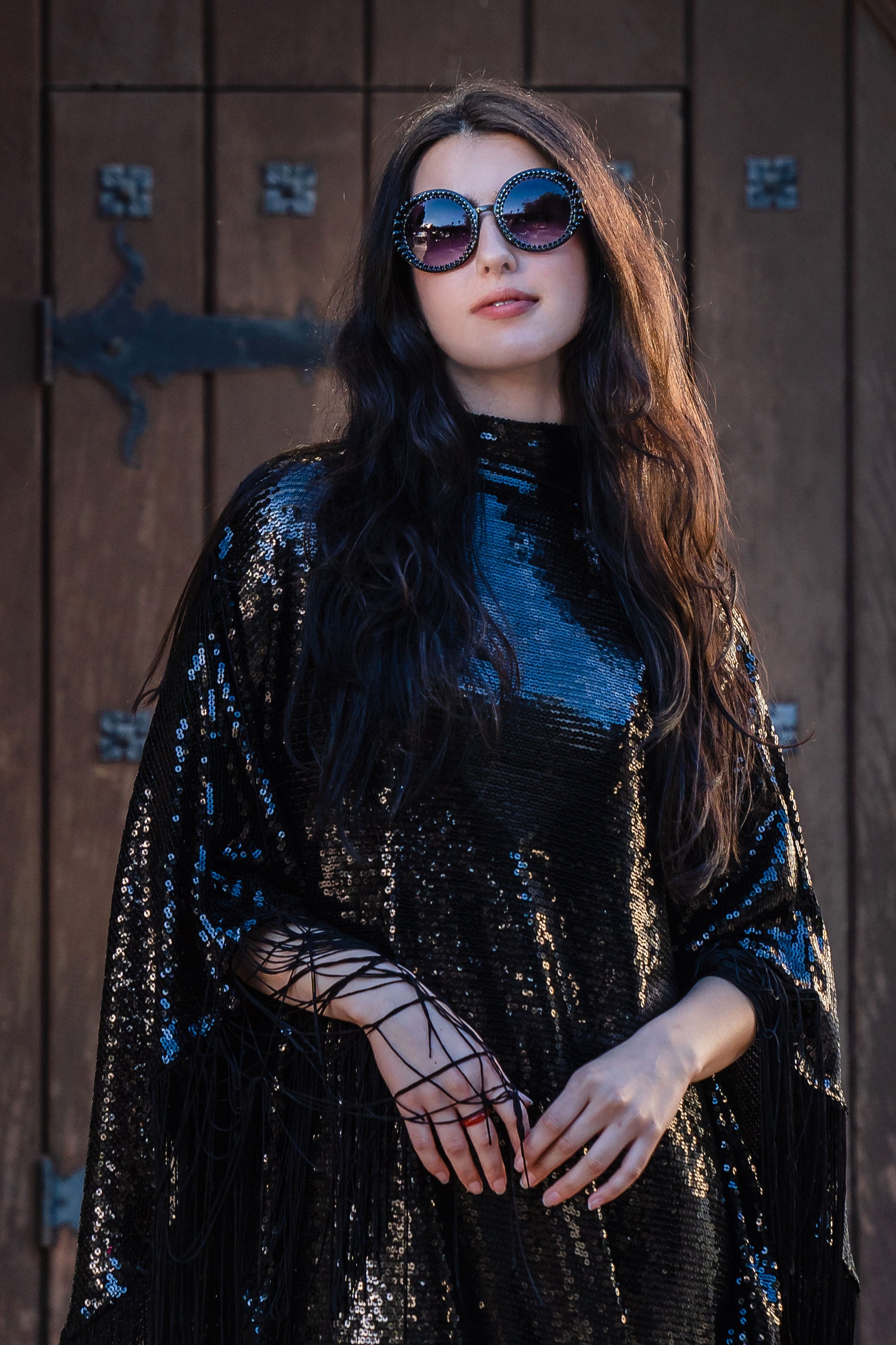 This caftan is a dazzling, full-length garment featuring a shimmering inky black sequin overlay on a mesh fabric. The sequins catch the light, creating a sparkling effect throughout. It has a mock neck collar with a straight, loose fit that drapes elegantly over the body. The edges of the caftan, including the hem and the sides, are embellished with long, black silk fringe that add movement and a playful flair to an elegant and timeless design.