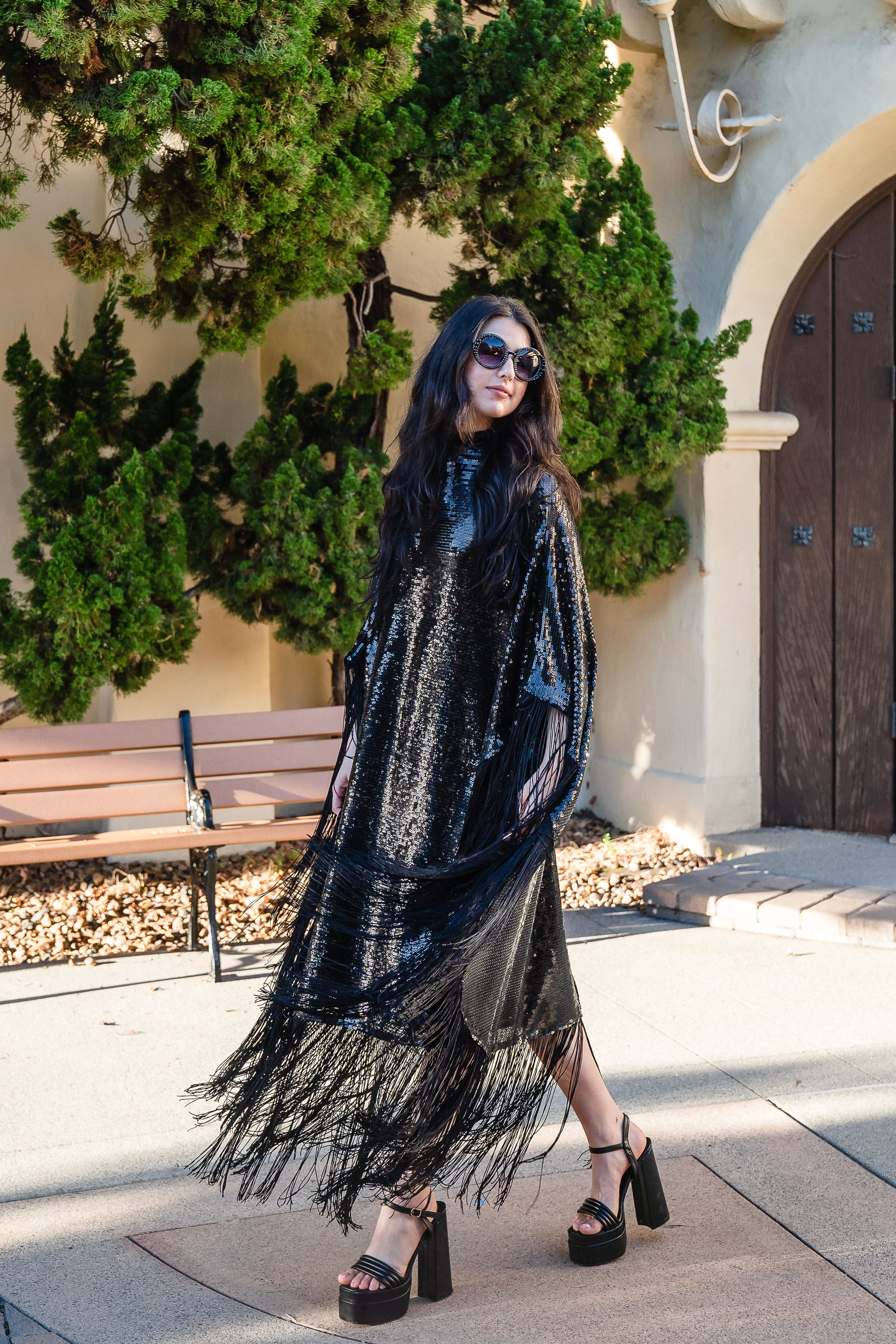 This caftan is a dazzling, full-length garment featuring a shimmering inky black sequin overlay on a mesh fabric. The sequins catch the light, creating a sparkling effect throughout. It has a mock neck collar with a straight, loose fit that drapes elegantly over the body. The edges of the caftan, including the hem and the sides, are embellished with long, black silk fringe that add movement and a playful flair to an elegant and timeless design.