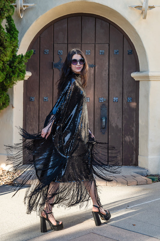 This caftan is a dazzling, full-length garment featuring a shimmering inky black sequin overlay on a mesh fabric. The sequins catch the light, creating a sparkling effect throughout. It has a mock neck collar with a straight, loose fit that drapes elegantly over the body. The edges of the caftan, including the hem and the sides, are embellished with long, black silk fringe that add movement and a playful flair to an elegant and timeless design.