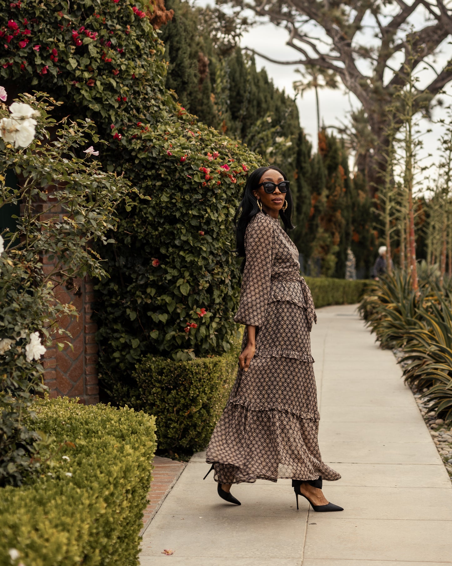 This beige and black floral maxi dress is crafted with a soft, metallic infused fabric, it features high neck, bishop sleeves, tiered ruffle skirt and matching sash for a cinched waist.