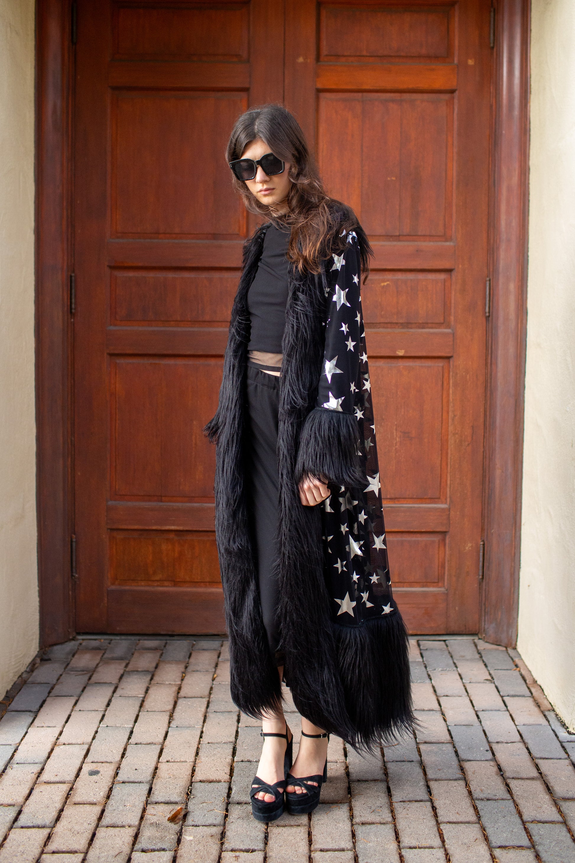Woman wearing a black outfit with a star-patterned coat in front of a wooden door.