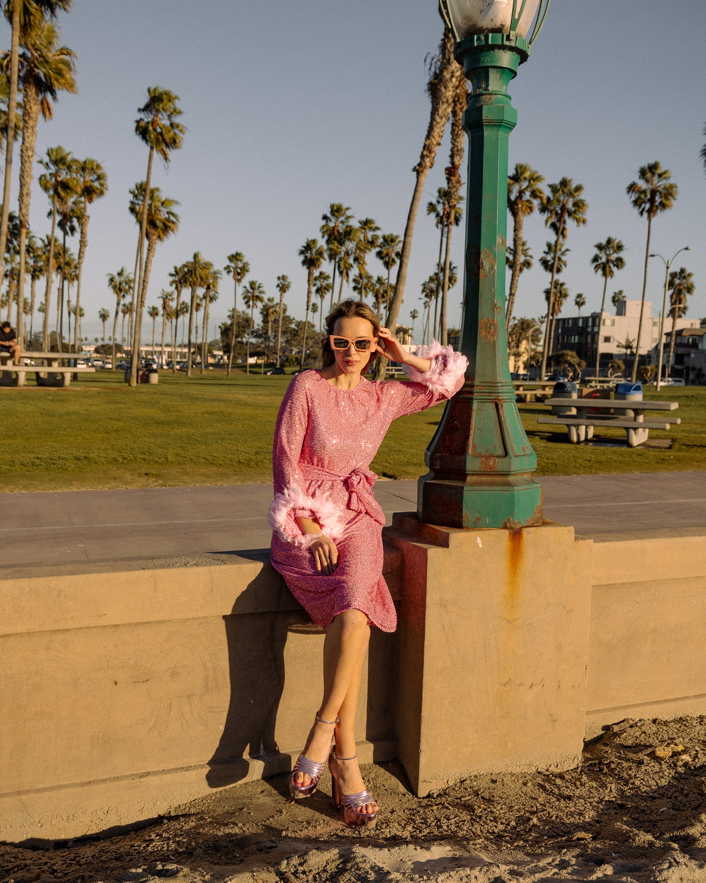 Jennafer Grace light pink flamingo sequins dress. Featuring a boat neck, generous bishop sleeves with light pink feather detail at sleeve hem, flowing midi length maxi skirt, and a slim matching sash for a cinched waist. 