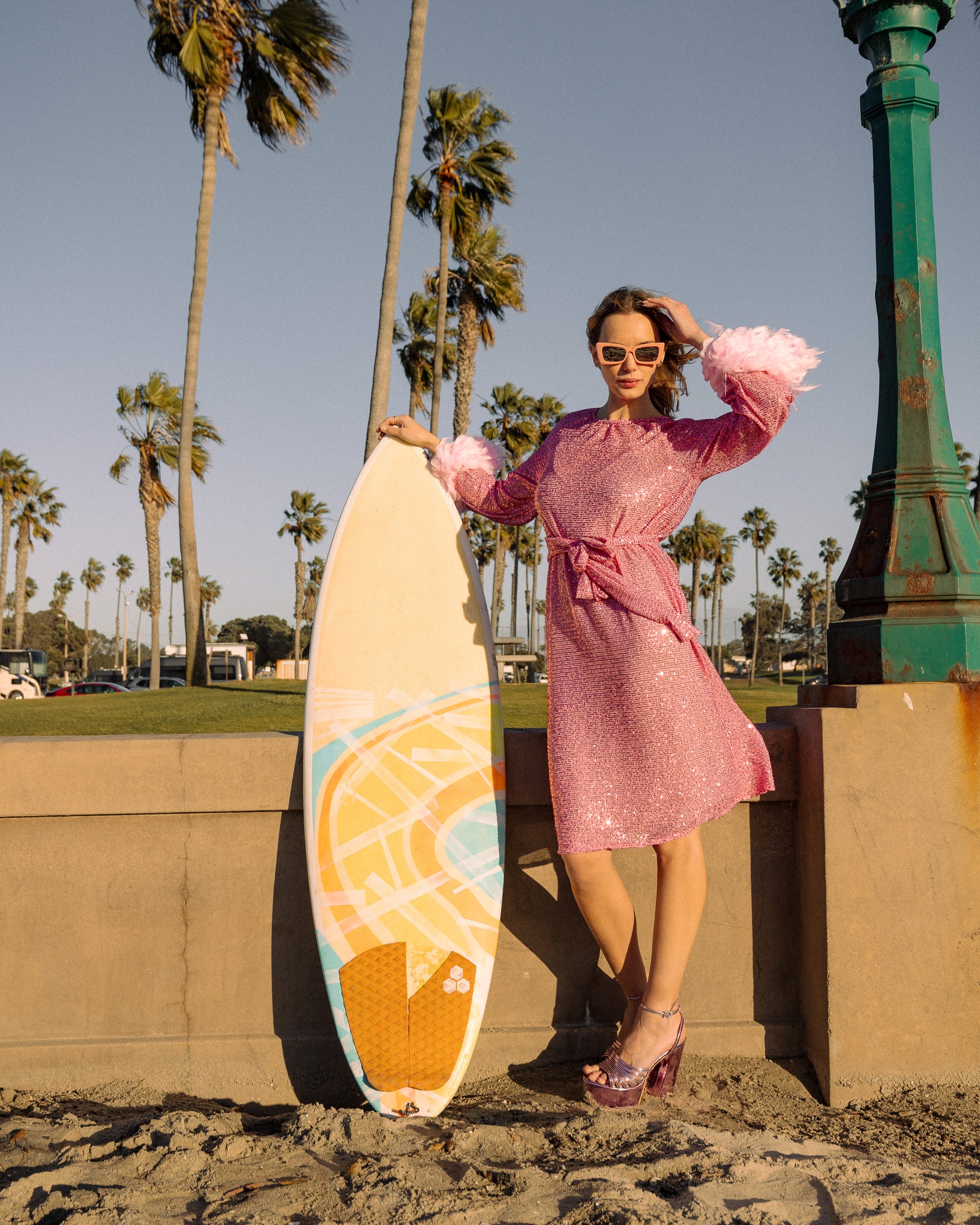 Jennafer Grace light pink flamingo sequins dress. Featuring a boat neck, generous bishop sleeves with light pink feather detail at sleeve hem, flowing midi length maxi skirt, and a slim matching sash for a cinched waist. 