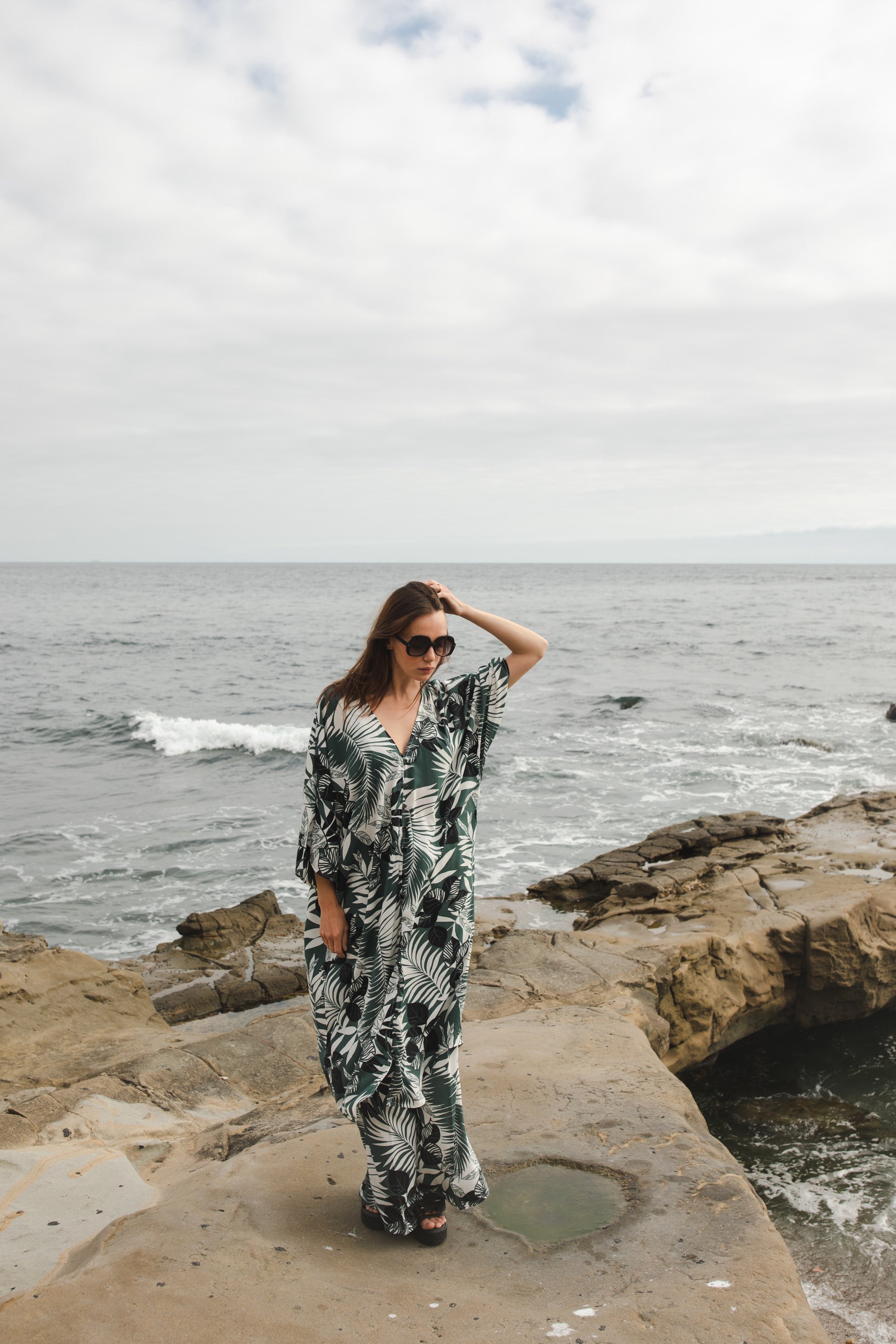 Midi length caftan tunic featuring white palm leaf print on dark hunter green. With v-neck, button up front, cutout at upper back with shoulder tie, and matching palazzo pant. Tropical bohemian chic aesthetic.