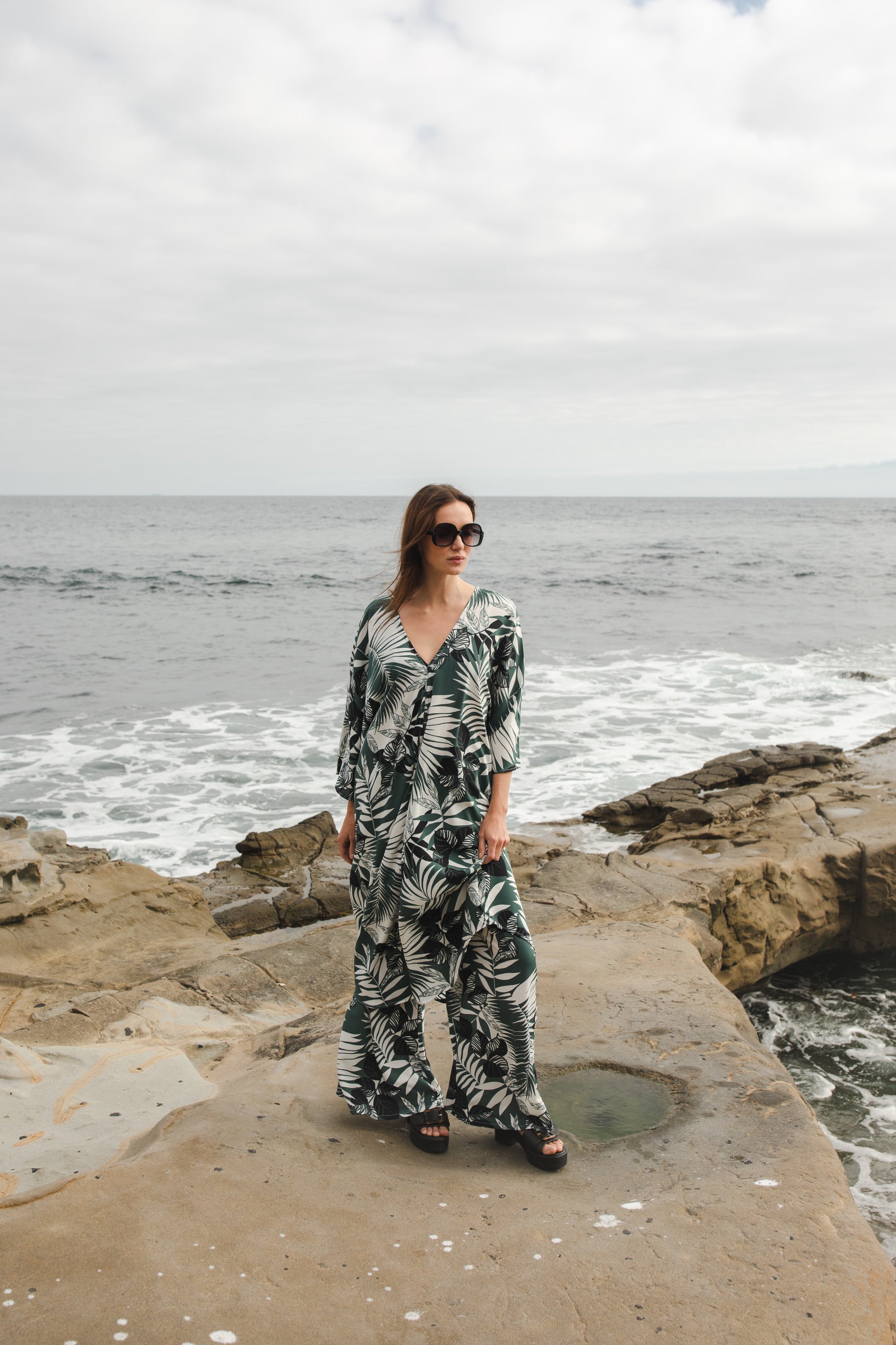 Midi length caftan tunic featuring white palm leaf print on dark hunter green. With v-neck, button up front, cutout at upper back with shoulder tie, and matching palazzo pant. Tropical bohemian chic aesthetic.