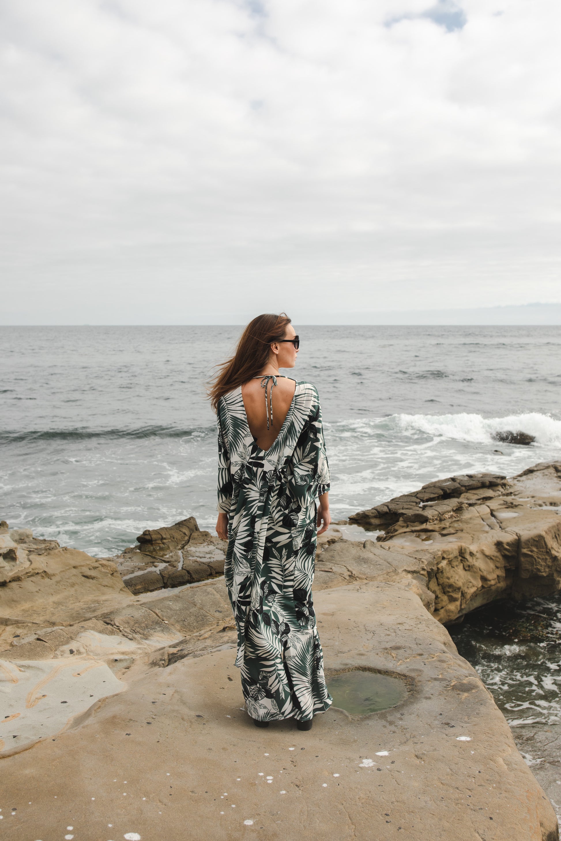 Midi length caftan tunic featuring white palm leaf print on dark hunter green. With v-neck, button up front, cutout at upper back with shoulder tie, and matching palazzo pant. Tropical bohemian chic aesthetic.