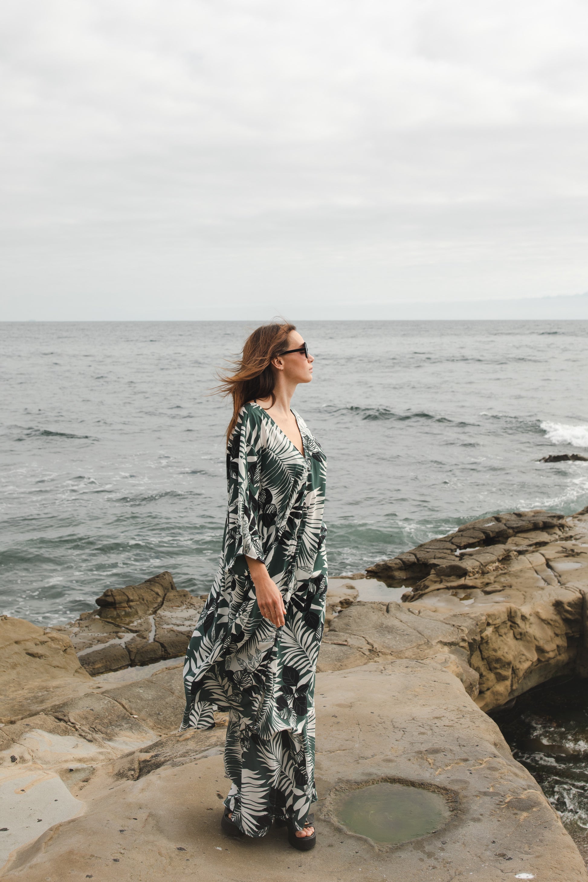 Midi length caftan tunic featuring white palm leaf print on dark hunter green. With v-neck, button up front, cutout at upper back with shoulder tie, and matching palazzo pant. Tropical bohemian chic aesthetic.