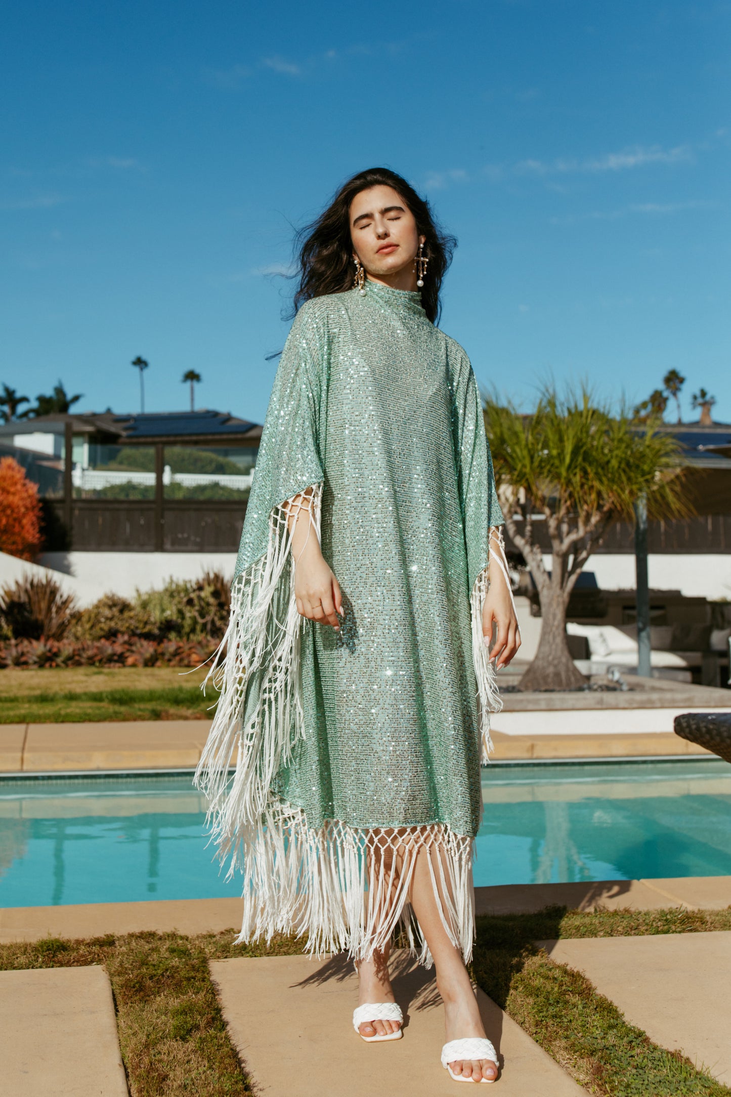 This caftan is a dazzling, full-length garment featuring a shimmering seafoam green sequin overlay on a mesh fabric. The sequins catch the light, creating a sparkling effect throughout. It has a mock neck collar with a straight, loose fit that drapes elegantly over the body. The edges of the caftan, including the hem and the sides, are embellished with long, ivory silk fringe that add movement and a playful flair to an elegant and timeless design.