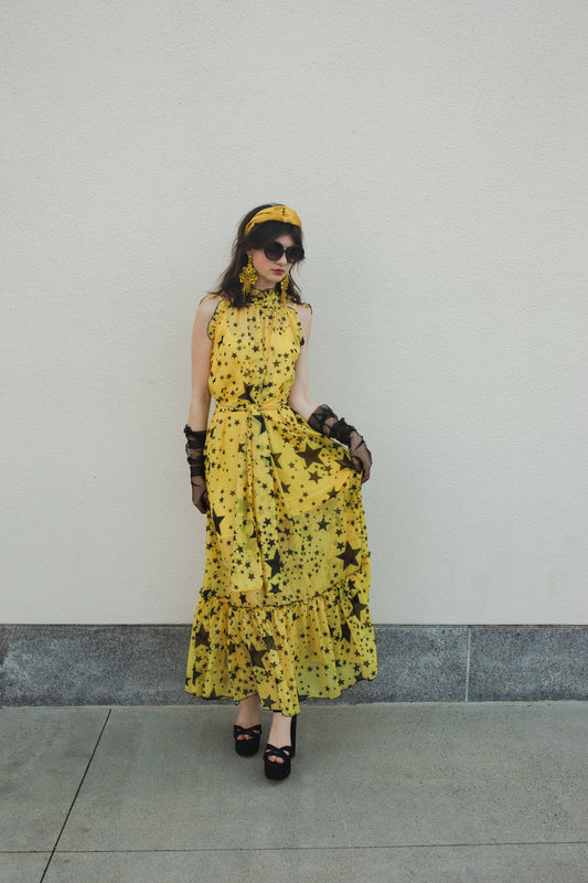 A maxi length a-line dress with ruffle at sleeve and hem, neck, and arm holes. Sleeveless with mockneck and ankle length hem. Yellow fabric with black stars in varying size. Comes yellow slip and matching waist tie.
