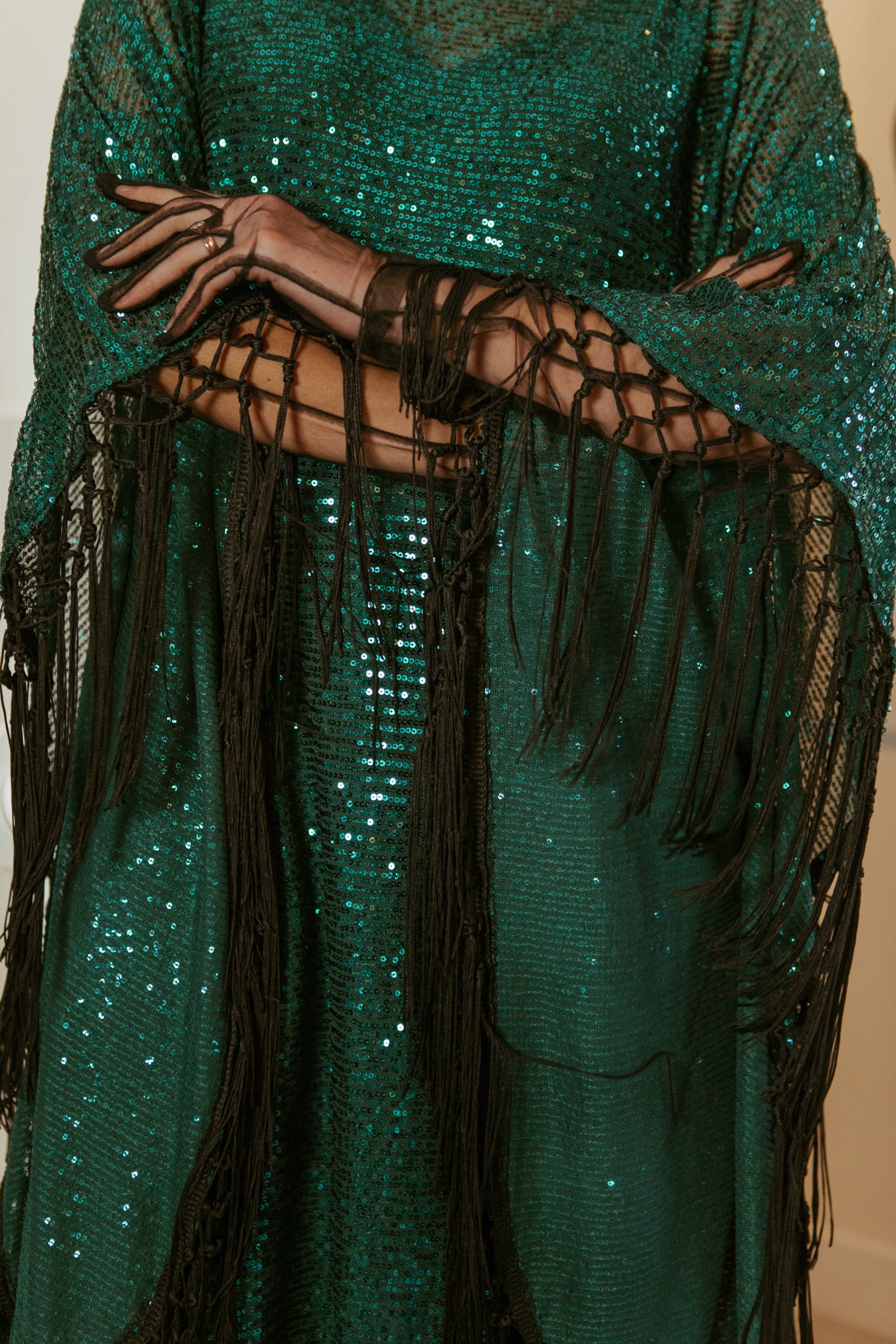 This caftan is a dazzling, full-length garment featuring a shimmering jade green sequin overlay on a mesh fabric. The sequins catch the light, creating a sparkling effect throughout. It has a mock neck collar with a straight, loose fit that drapes elegantly over the body. The edges of the caftan, including the hem and the sides, are embellished with long, silk black fringe that add movement and a playful flair.